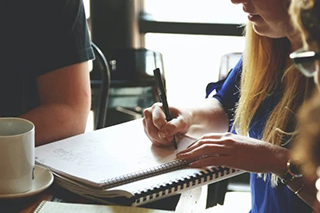 Woman writing notes on lined paper at a coffee shop with other team members